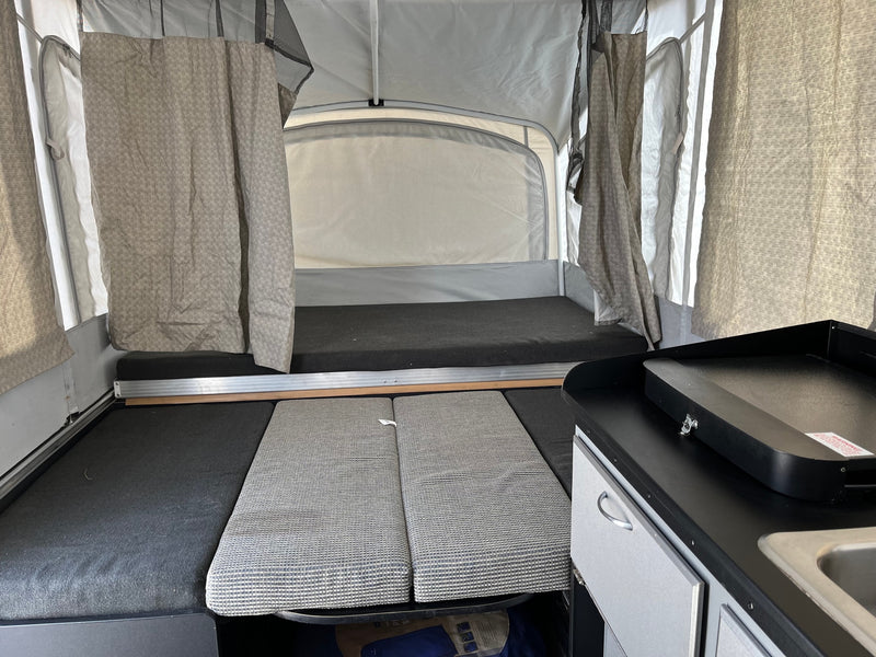 Inside an RV rental pop-up camper: a view of the bed, kitchen counter with griddle, and seating cushions. — photo 11
