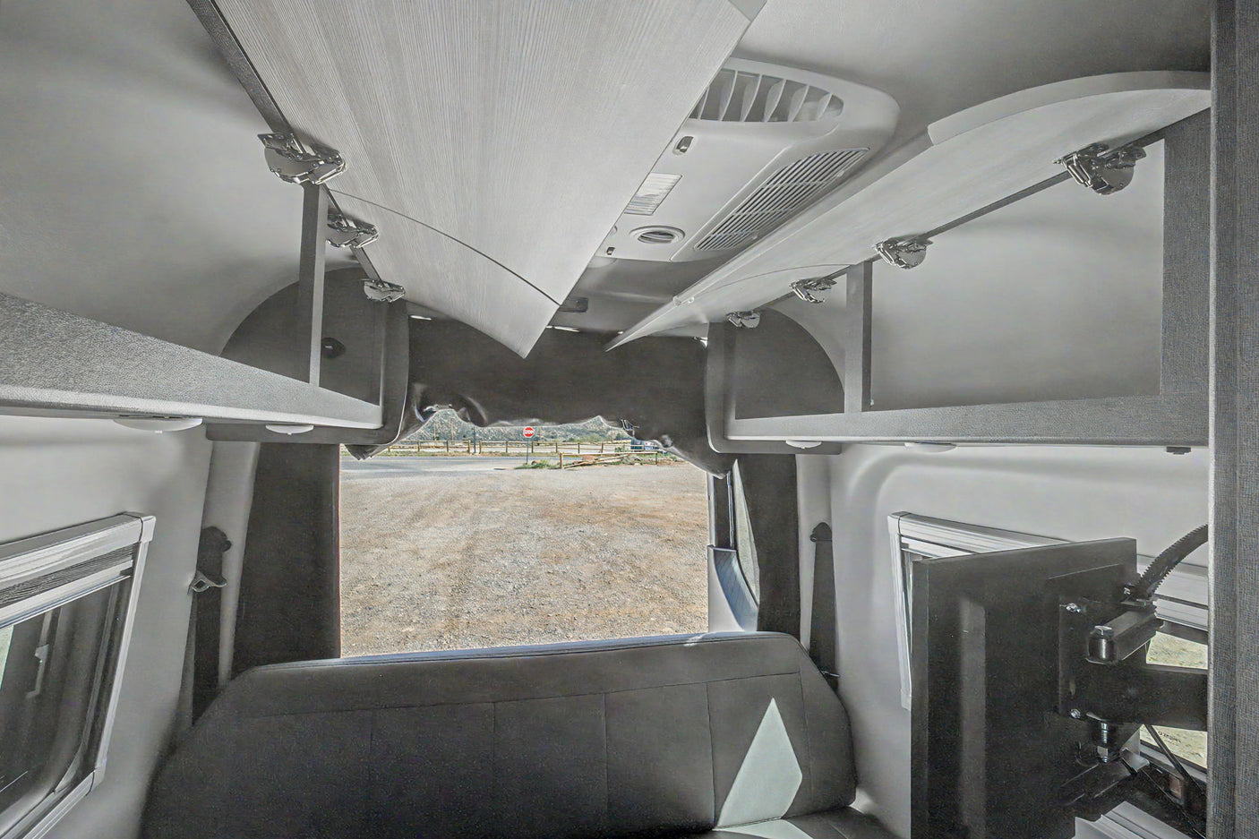 RV rental interior: open overhead storage, rear bench seat, TV, and view of a gravel road and hills out the back.