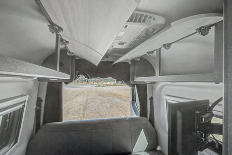 RV rental interior: open overhead storage, rear bench seat, TV, and view of a gravel road and hills out the back.