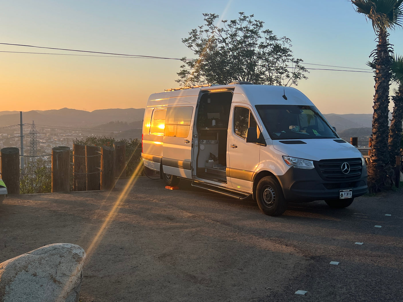 White Sprinter RV rental parked at a scenic sunset. Side door open, sunlight reflecting off the van, mountains in the distance.
