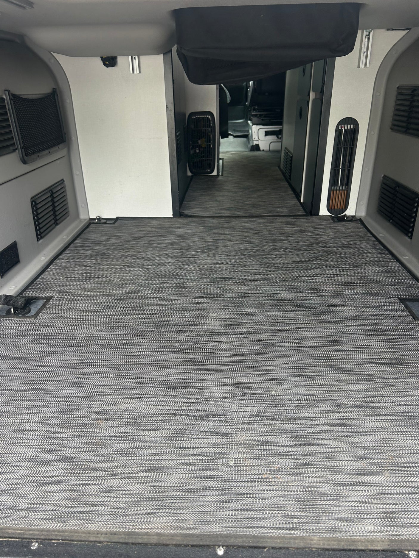 RV rental interior: Spacious cargo/living area with gray textured flooring, side panels, vents, and tie-downs, leading to the cab.