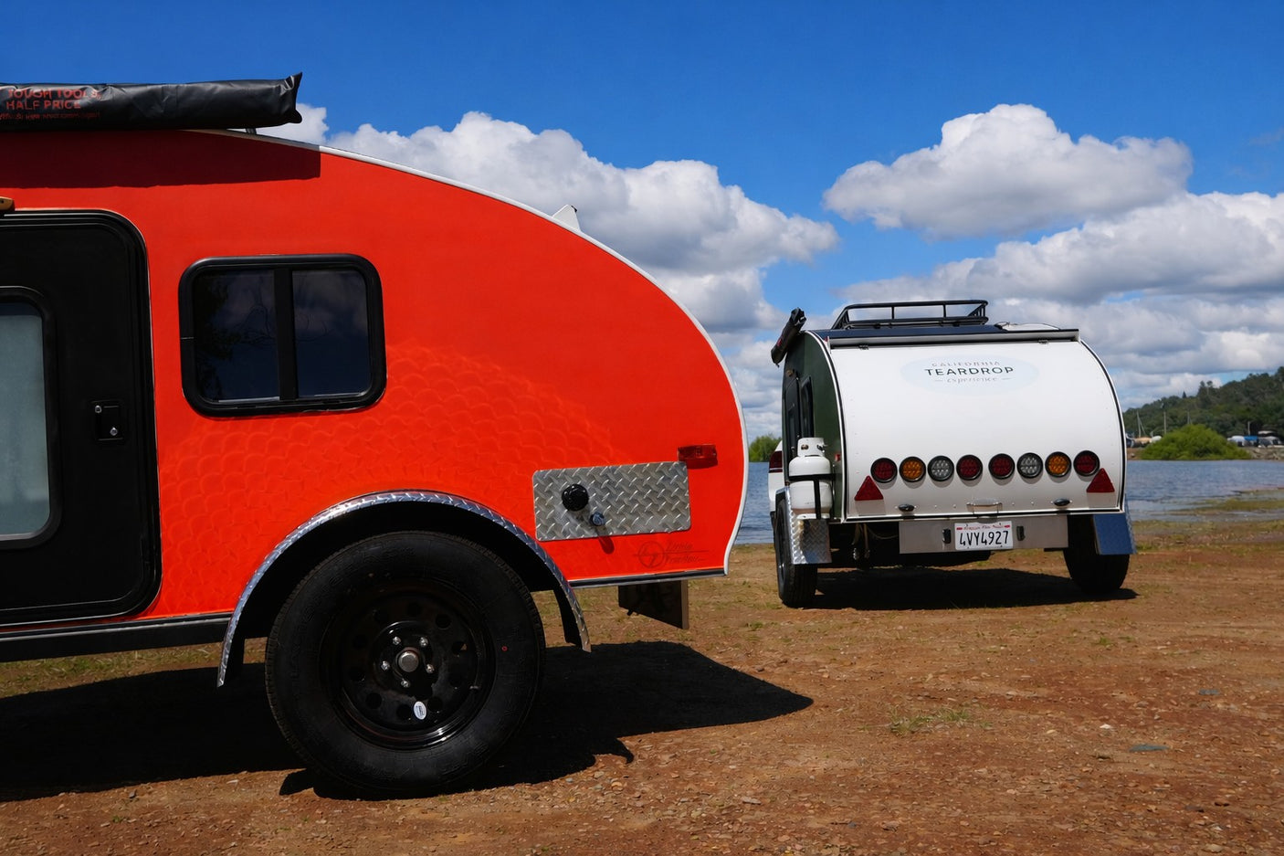Bright red and white teardrop RV rental campers are parked by a scenic lake under a blue sky with fluffy clouds.