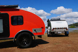 Bright red and white teardrop RV rental campers are parked by a scenic lake under a blue sky with fluffy clouds.