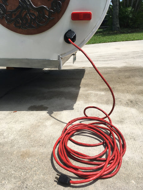 Hooking up an RV rental: Red extension cord provides shore power to a white camper on a sunny driveway.