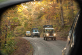 Fall foliage frames a convoy of classic 4x4s on a winding road, seen from a car's side mirror. Consider an RV rental for your next adventure!