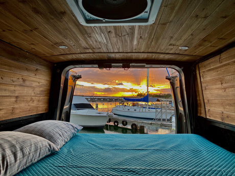 Cozy rv rental bed looking out to a stunning orange sunset over a marina filled with boats. Perfect for van life adventures.