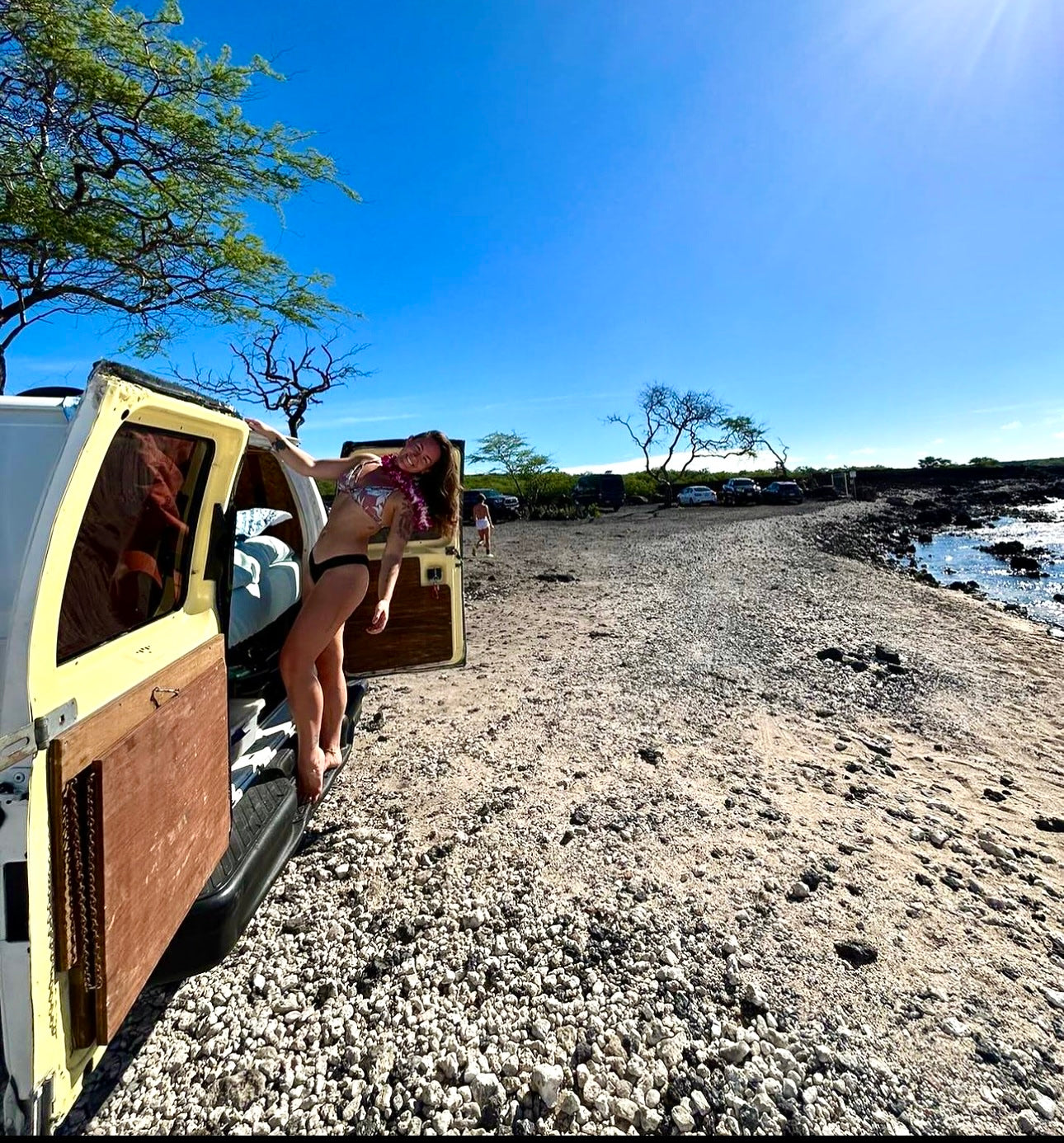 Sunny beach adventures in an RV rental! A woman in a bikini and lei smiles, leaning out of her camper van on a rocky shore.