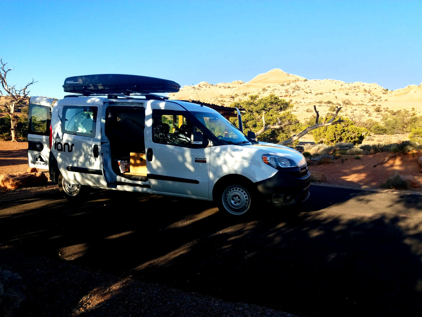Explore nature with this white Ram ProMaster City RV rental van, doors open, parked amidst desert scenery under a clear sky.