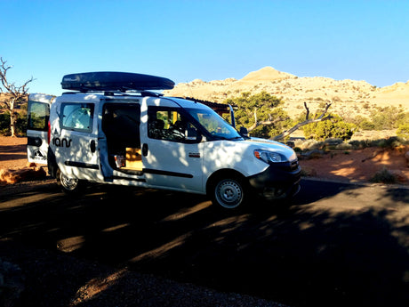 Explore nature with this white Ram ProMaster City RV rental van, doors open, parked amidst desert scenery under a clear sky.