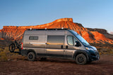Gray RV rental van with mountain bike on a dirt road, facing sunlit red mountains under a blue sky. Perfect for adventure.