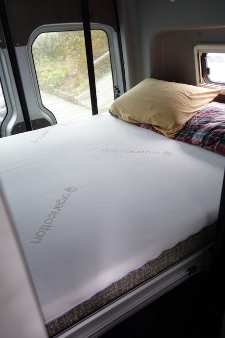 RV rental's cozy bed: a white organiCotton mattress, pillow, and plaid blanket inside a camper van, with window views.