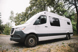 White camper van for RV rental parked on a gravel path, ready for outdoor adventures amidst lush trees.