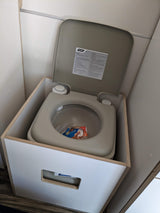 An RV rental Camco portable toilet, lid open showing instructions, integrated into a compact wooden enclosure.