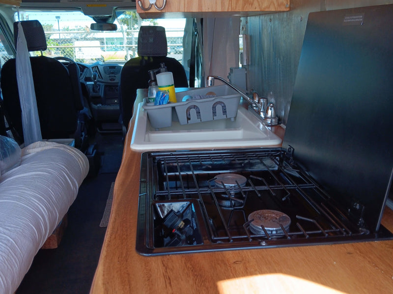 RV rental interior: A compact camper van kitchen with sink, faucet, and two-burner stove. Front cab also visible. — photo 10