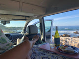 A person relaxes in an RV rental, feet up, enjoying wine, snacks, and ocean views from inside their campervan.