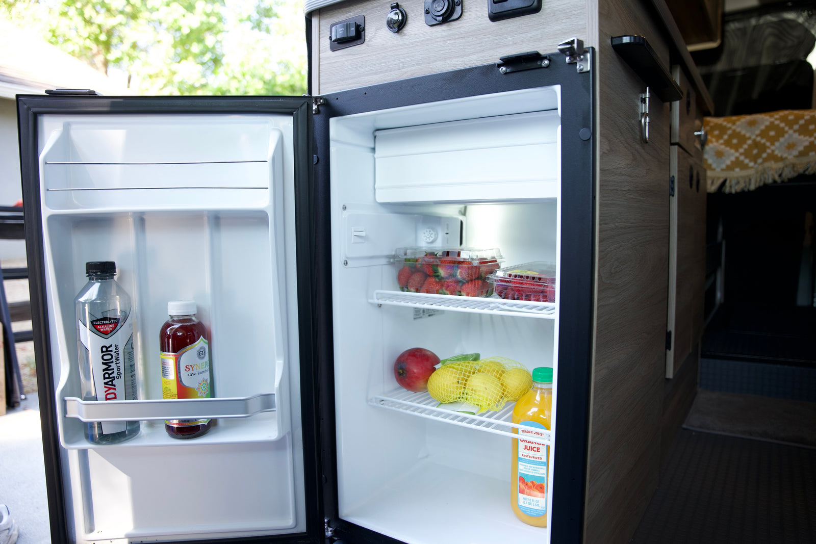 An open RV rental fridge, neatly packed with fresh fruit, kombucha, and juice. Ready for your next adventure!