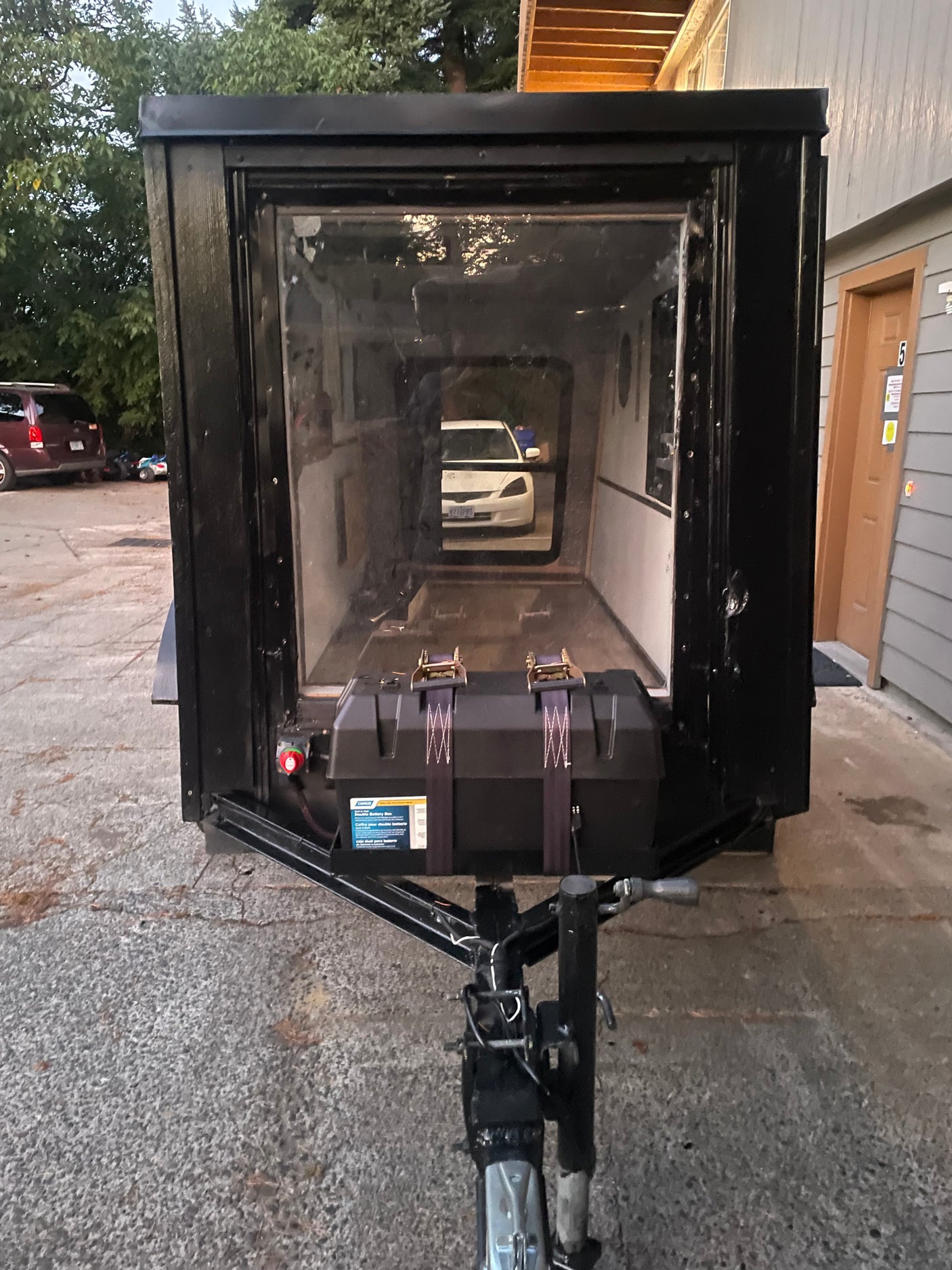 Compact black RV rental camper trailer with a transparent front panel, external battery box, and hitch, parked outdoors.