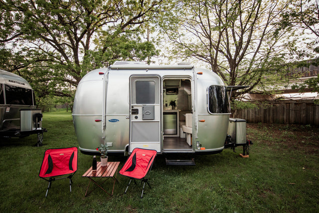 Silver Airstream rv rental with open door showing its interior, plus red chairs & a table outside in a grassy, treed setting.