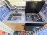 Compact RV rental kitchen: stainless sink, Dometic stove, fridge, and vibrant patterned tile backsplash under a window.