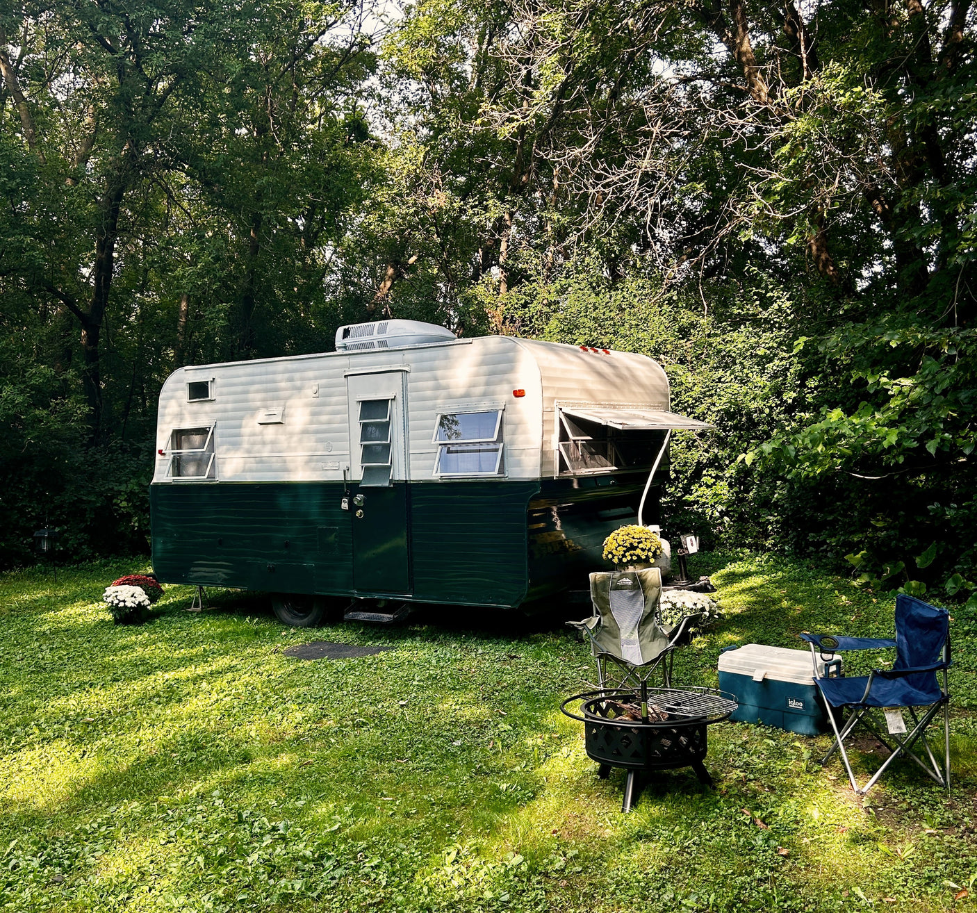 Cozy vintage RV rental in a lush green campsite with chairs, cooler, and fire pit under tall trees.