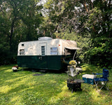 Cozy vintage RV rental in a lush green campsite with chairs, cooler, and fire pit under tall trees.