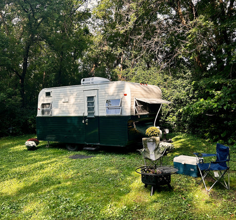 Cozy vintage RV rental in a lush green campsite with chairs, cooler, and fire pit under tall trees.