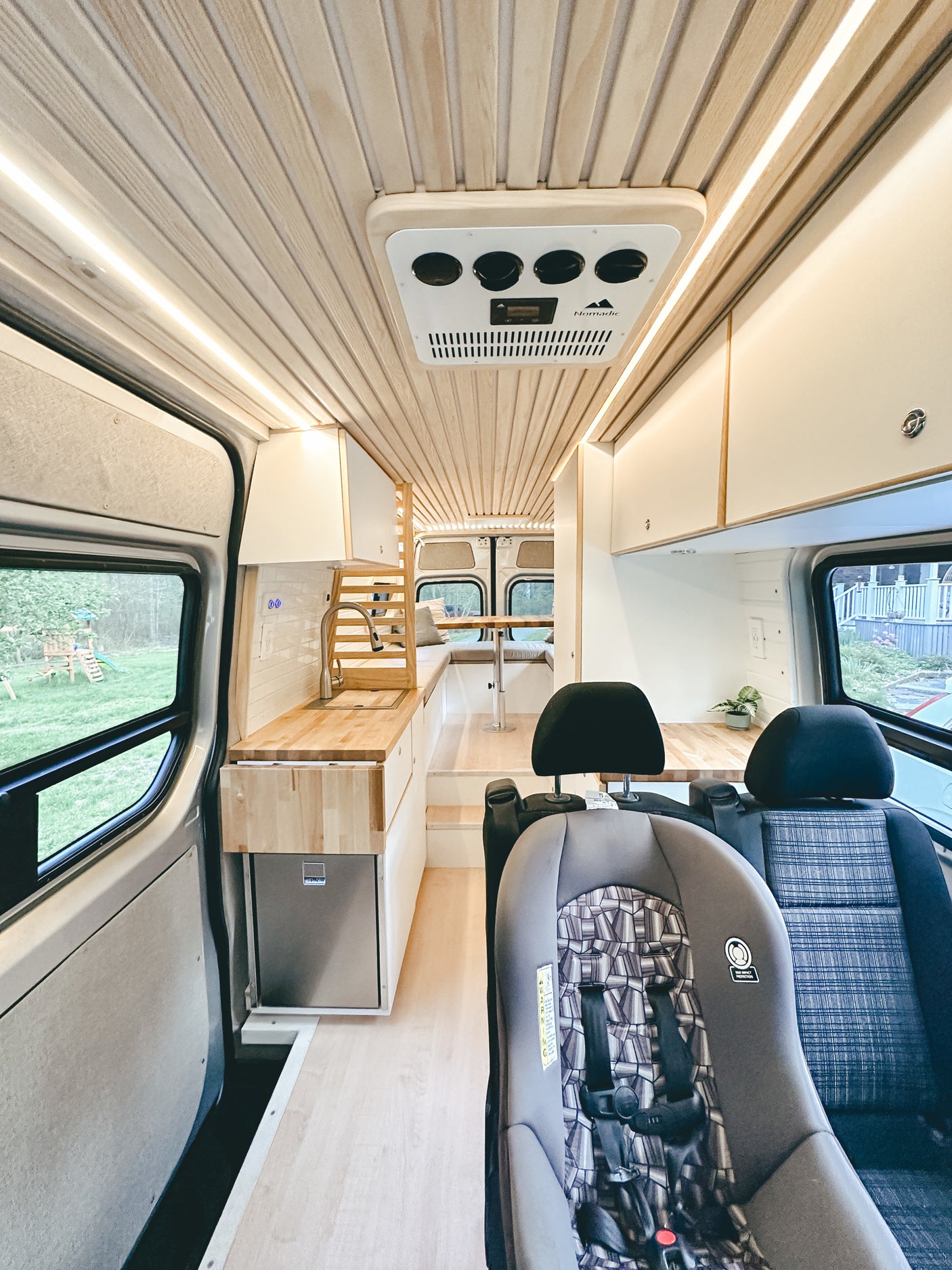 Bright RV rental interior: wood-paneled ceiling, modern kitchen with sink, fridge, and cozy seating. Child car seat in front.
