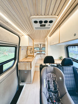 Bright RV rental interior: wood-paneled ceiling, modern kitchen with sink, fridge, and cozy seating. Child car seat in front.