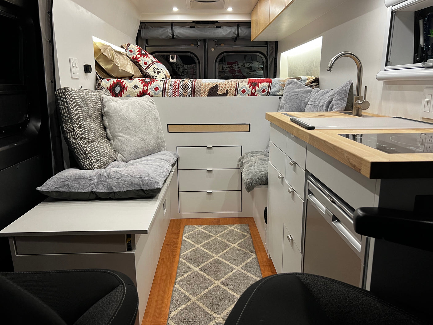 Inside a comfy rv rental, featuring a bed with patterned quilt, compact kitchen, and a plush seating area with storage.