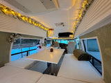 Step into a festive RV rental! This cozy camper features lit garland, a table, and plush seating, with snow outside.