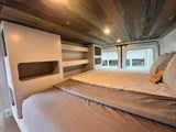 RV rental: Inside a comfortable camper van bedroom with a bed, light bedding, wood-paneled ceiling, and shelving.
