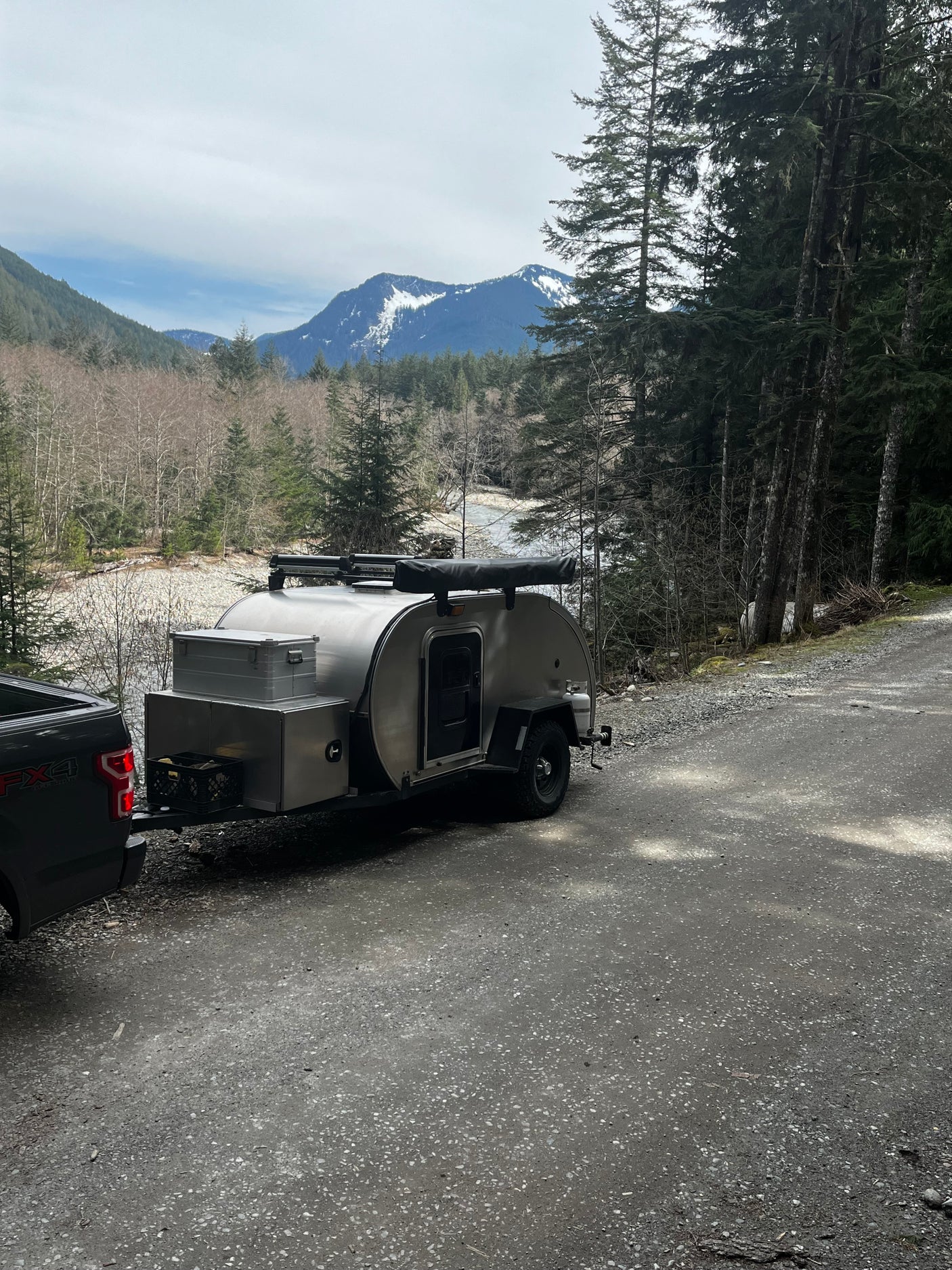 A silver teardrop camper and truck are parked by a scenic mountain river. Ideal for an RV rental adventure.