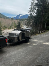 A silver teardrop camper and truck are parked by a scenic mountain river. Ideal for an RV rental adventure.