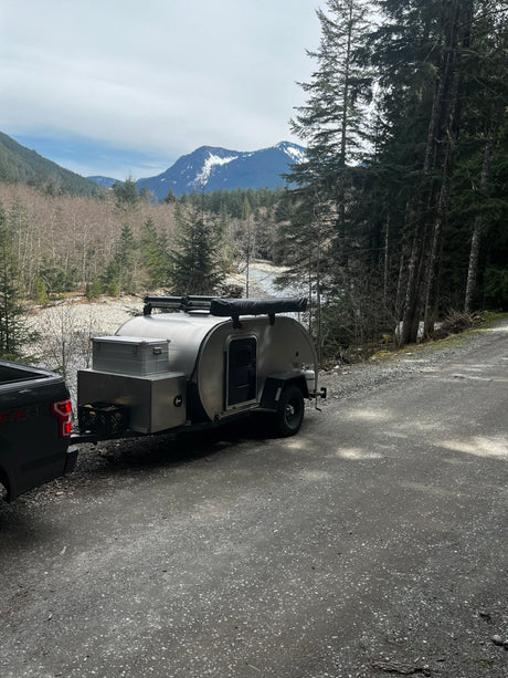 A silver teardrop camper and truck are parked by a scenic mountain river. Ideal for an RV rental adventure.