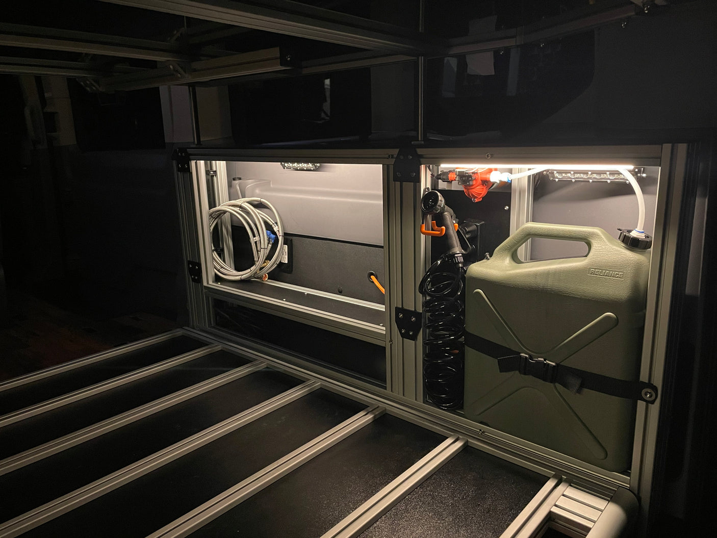 Compact water system in an RV rental: Features an illuminated storage area with a pump, shower head, and portable water tank.