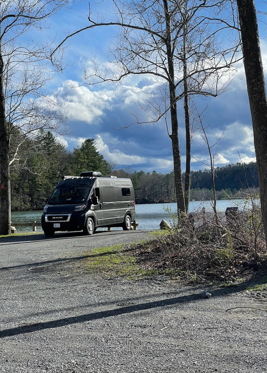 Black RV rental parked on a gravel road beside a serene lake, framed by bare trees, under dramatic clouds. Ideal for nature trips.