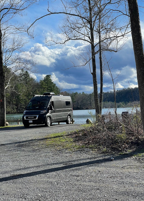 Black RV rental parked on a gravel road beside a serene lake, framed by bare trees, under dramatic clouds. Ideal for nature trips.