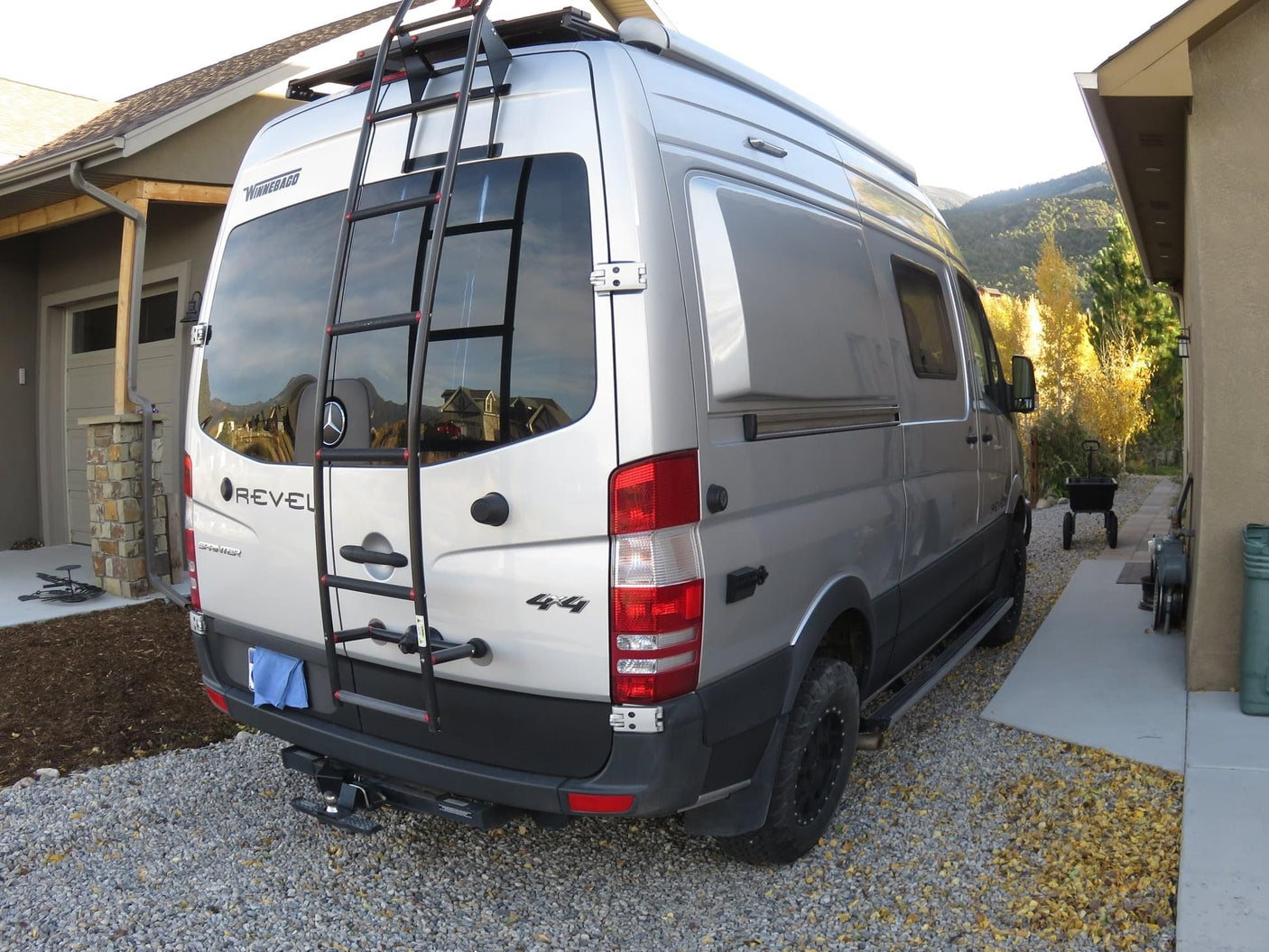 Silver Winnebago Revel 4x4 camper van, featuring a rear ladder, on a gravel driveway. Ideal for RV rental adventures.