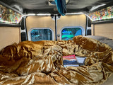 A plush gold blanket on a bed inside a cozy rv rental van, overlooking a serene campsite with tents. Perfect for travel adventure.