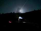Experience starry nights: a moonlit camping vehicle with tent (like an RV rental) under a plane trail & glowing campfire.