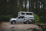 RV rental: A silver Toyota Tacoma with an extended pop-up truck camper, parked on a gravel forest road amidst tall trees.