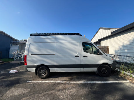 White Mercedes Sprinter van with roof rack, parked on asphalt. Excellent choice for an RV rental.