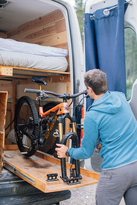A man loads his orange Rocky Mountain bike onto a pull-out tray in the back of an RV rental van.