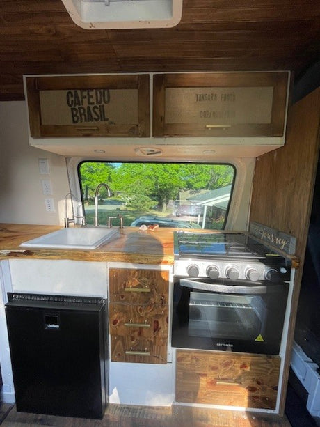 RV rental interior showcases a functional kitchen with sink, compact fridge, stove/oven, and unique wood cabinetry.
