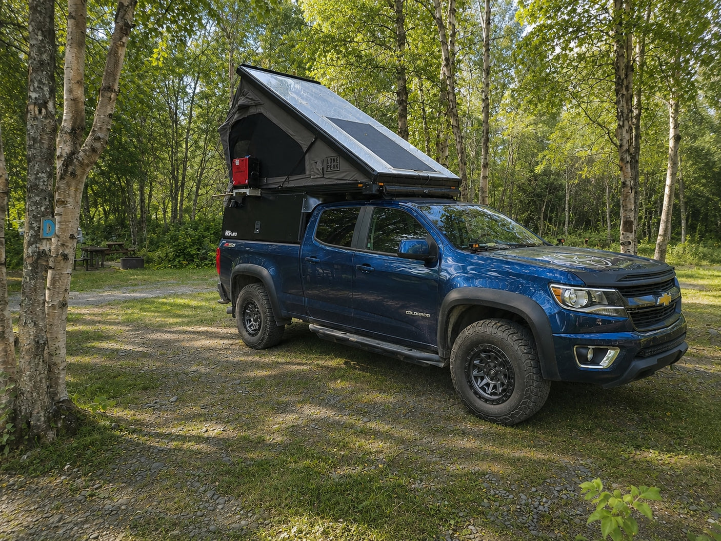 This blue Chevy Colorado with a rooftop tent and truck camper is parked at a shaded campsite, ready for an adventure RV rental.