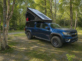 This blue Chevy Colorado with a rooftop tent and truck camper is parked at a shaded campsite, ready for an adventure RV rental.