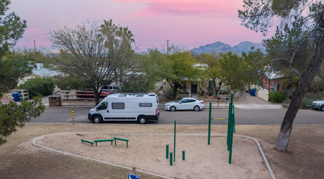 Scenic view of an rv rental parked on a street near a park with exercise equipment, houses, mountains & pink sky at sunset.