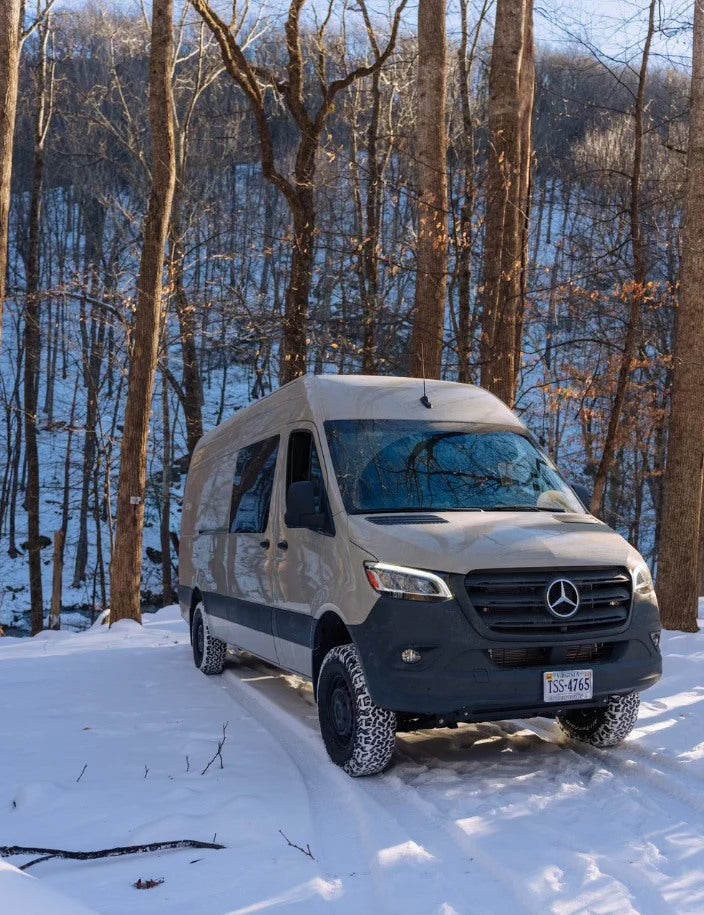 Beige Mercedes Sprinter RV rental van on snow, tire treads visible, parked in a winter forest. Ready for adventure.