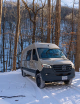 Beige Mercedes Sprinter RV rental van on snow, tire treads visible, parked in a winter forest. Ready for adventure.