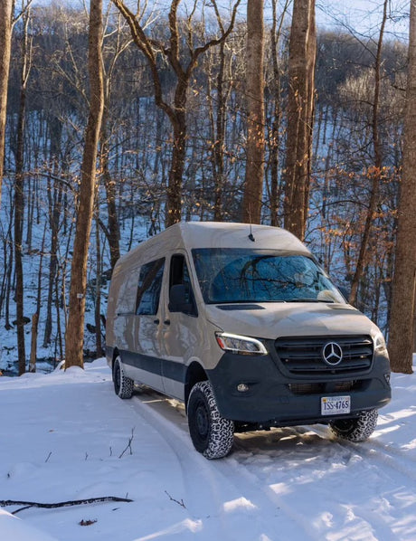 Beige Mercedes Sprinter RV rental van on snow, tire treads visible, parked in a winter forest. Ready for adventure.